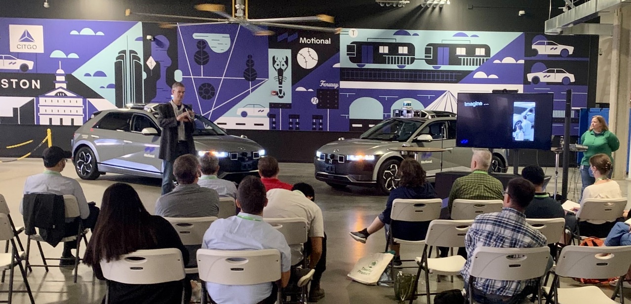 Motional's Paul Schmitt addresses a seated crowd while standing in front of two IONIQ 5 robotaxis