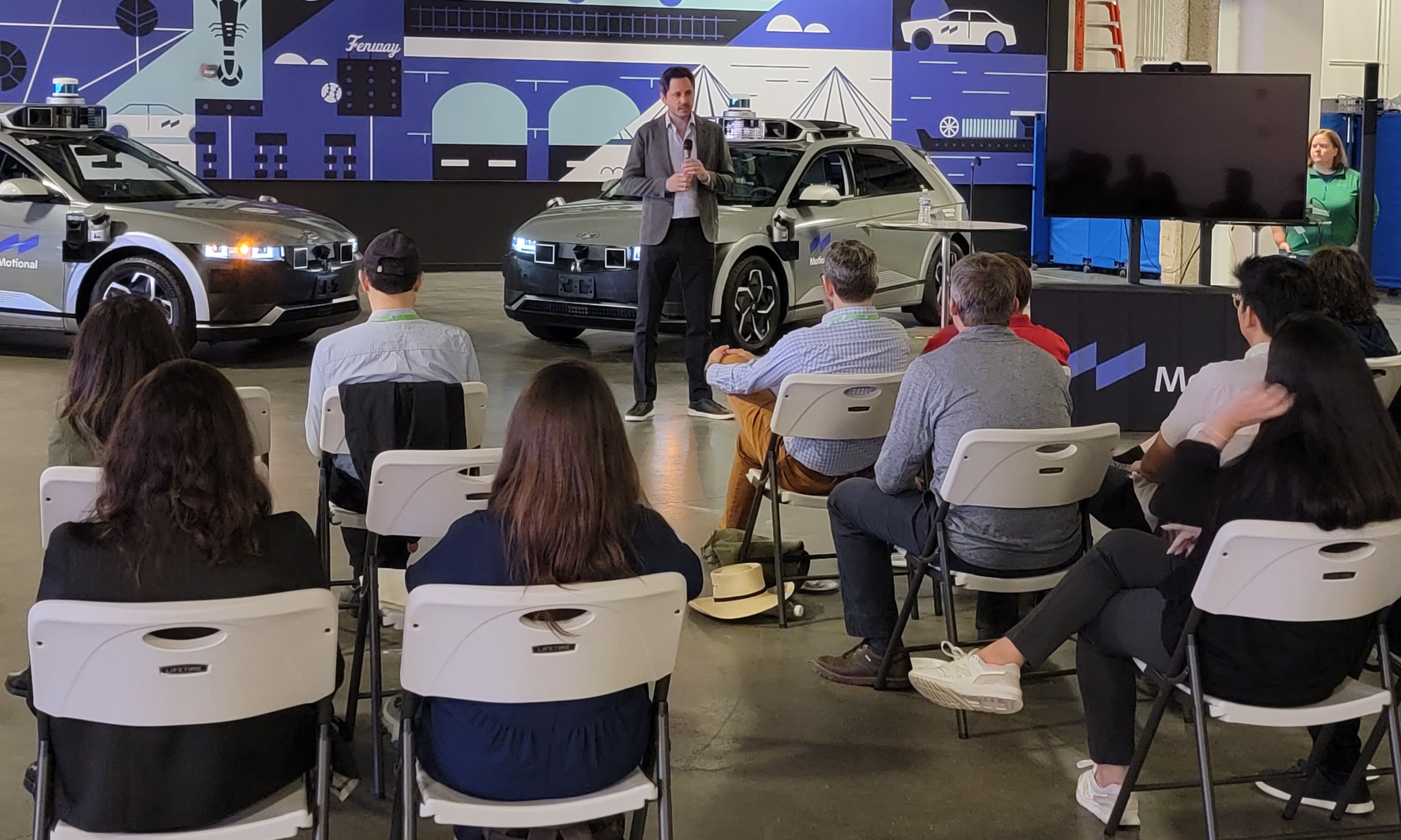 Motional President & CEO Karl Iagnemma stands in front of two IONIQ 5 robotaxis inside a garage addressing a seated crowd