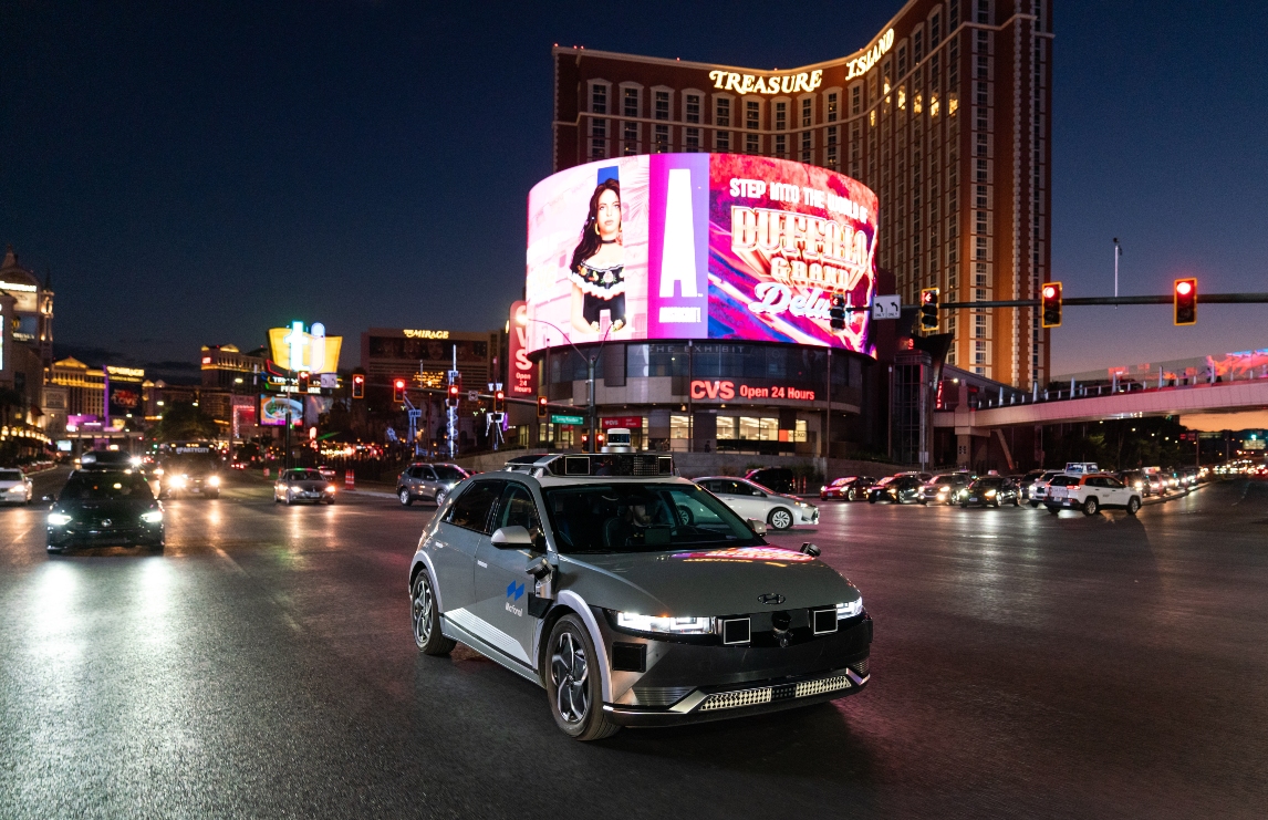 A gray IONIQ 5 robotaxi drives through a busy Las Vegas intersection just after dusk. A giant LED billboard is in the background. 