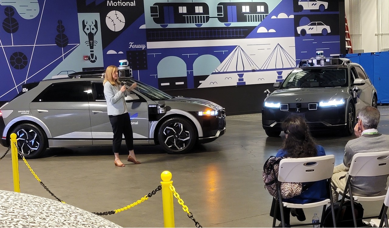 A woman stands in front of a gray IONIQ 5 robotaxi speaking to a seated audience