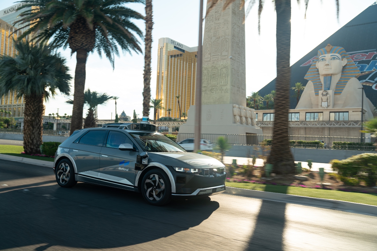 A gray IONIQ 5 robotaxi drives down the Las Vegas Strip during the daytime. There's palm trees on the median strip and a big casino in the background. 