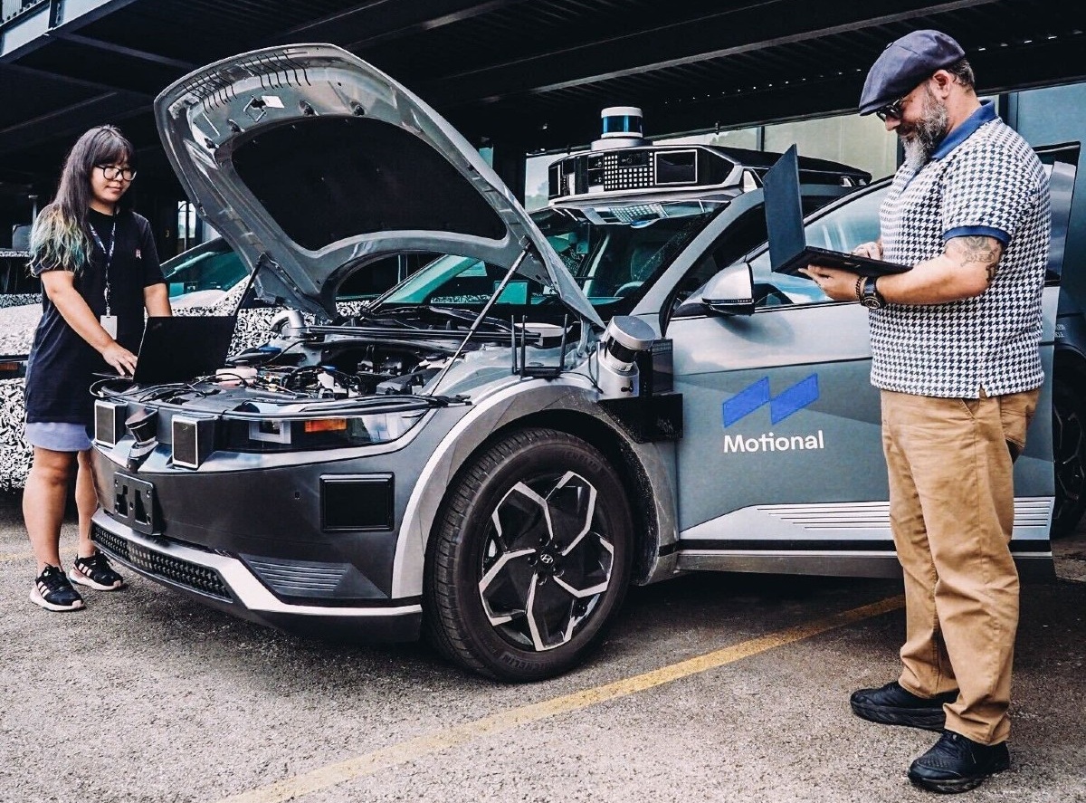A woman and man stand alongside an IONIQ 5 robotaxi. The vehicle's hood is up and the pair are conducting cybersecurity tests.