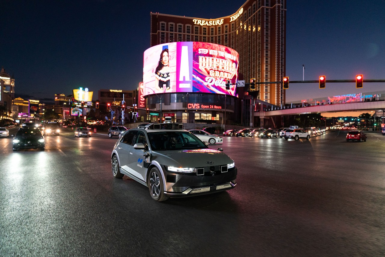 A silver Motional IONIQ 5 robotaxi drives through a busy Las Vegas intersection with a bright video screen in the background